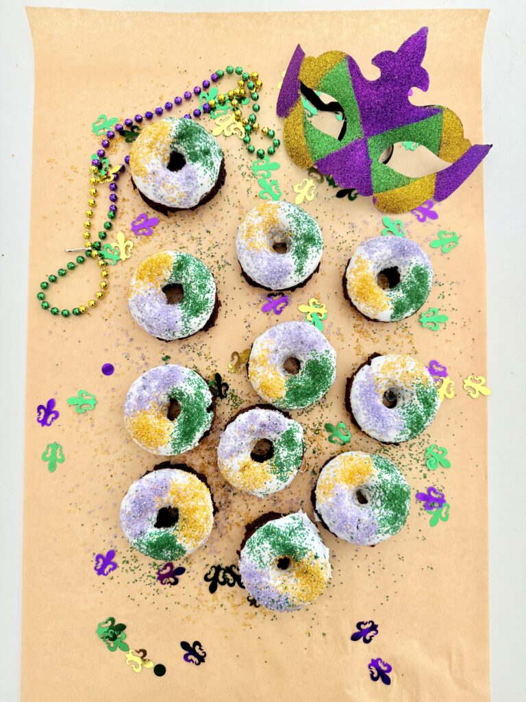 Mardi Gras donuts on brown parchment paper on the counter surrounded by mardi gras decorations.