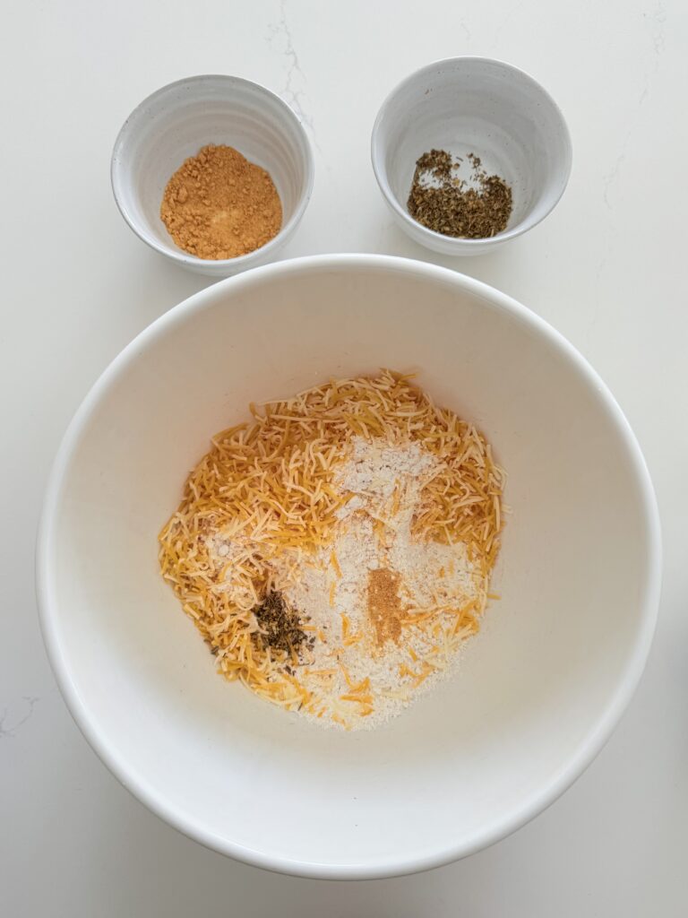 A large white mixing bowl filled with Bisquick mix, cheddar cheese, garlic powder, and Italian seasonings all on the counter.