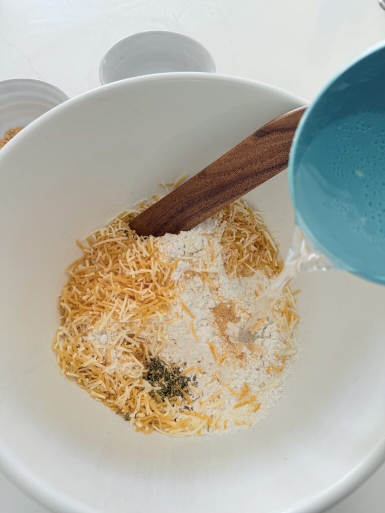 Pouring sparkling water into a large white mixing bowl filled with Bisquick mix, cheddar cheese, garlic powder, and Italian seasonings.