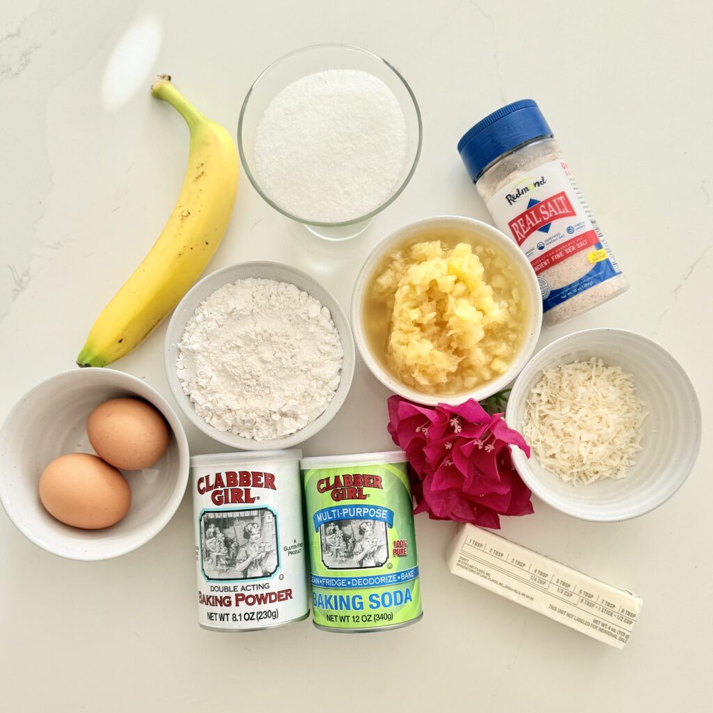 Flour in a white bowl, crushed pineapple in a white bowl, shredded coconut in a white bowl, two eggs in a white bowl, baking soda, baking powder, a stick of butter,, a ripe banana, sugar in a glass, salt, and bright pink flowers on the counter.