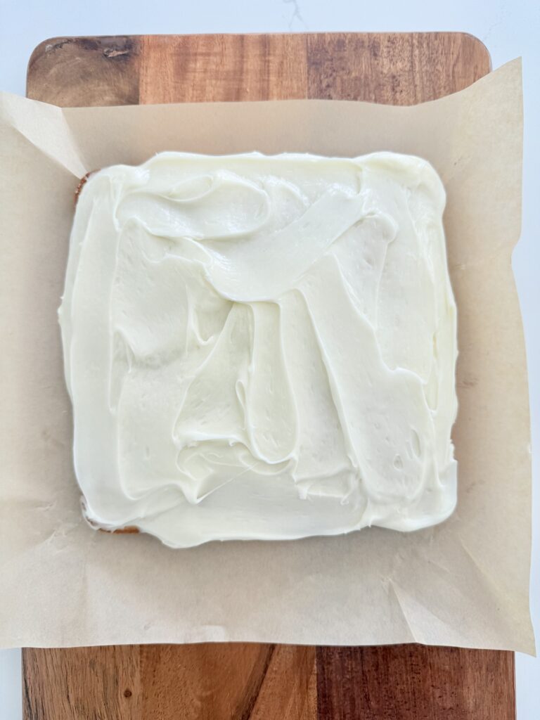 Frosted Almond Afternoon Cake on parchment paper on a brown cutting board.