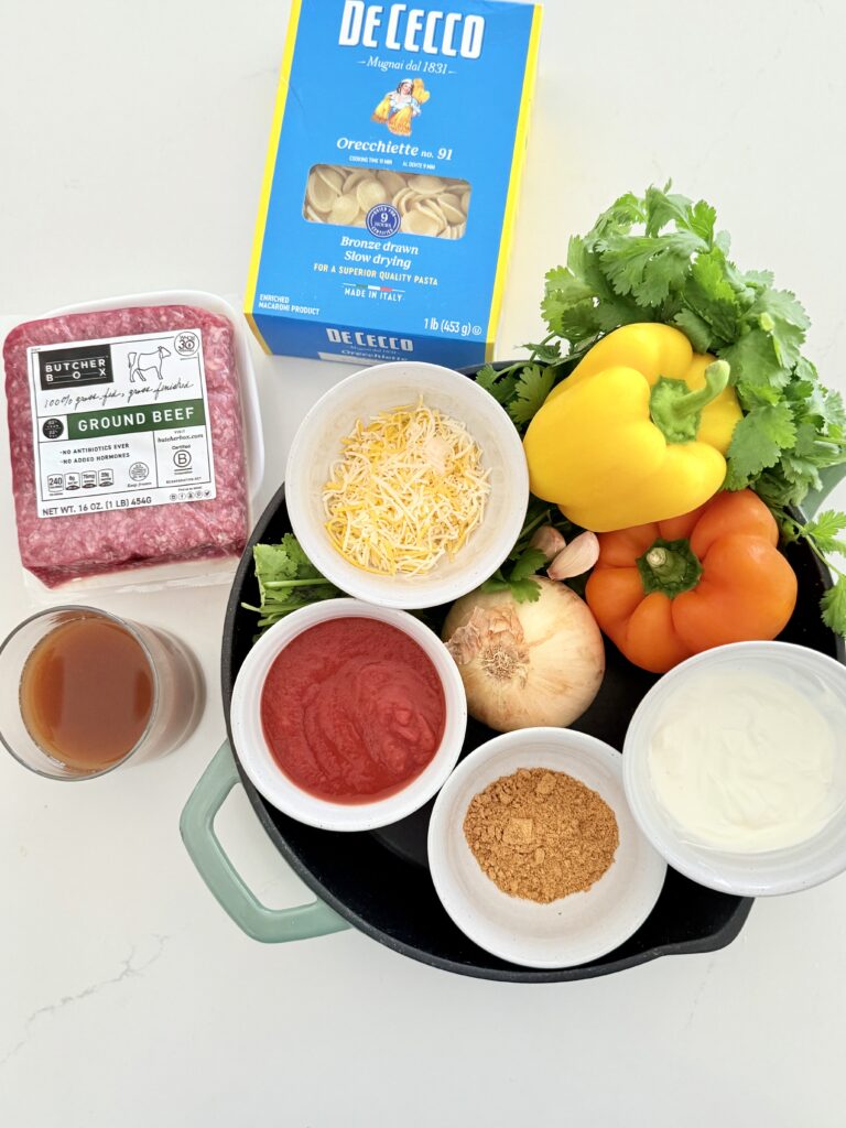 Lean ground beef, an onion, yellow and orange, bell peppers, garlic cloves, sour cream, and a white bowl, Mexican cheese blend in a small bowl, tomato sauce and a small bowl taco seasoning in a small bowl, beef broth in a glass, cilantro all in a cast-iron pan and the dried pasta on the counter.