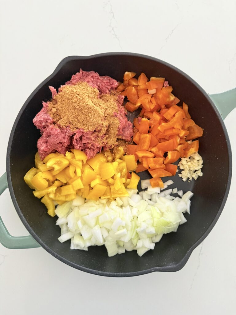 Ground beef with taco seasoning on top of it, red and yellow, diced bell peppers, diced onion, and minced garlic cloves, all in a cast-iron pan. On the counter.