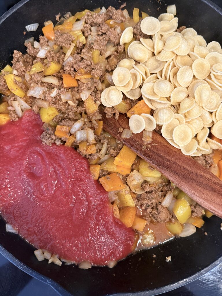 Ground beef with taco seasoning on top of it, red and yellow, diced bell peppers, diced onion, dried pasta, and minced garlic cloves, all in a cast-iron pan on the stove-top.