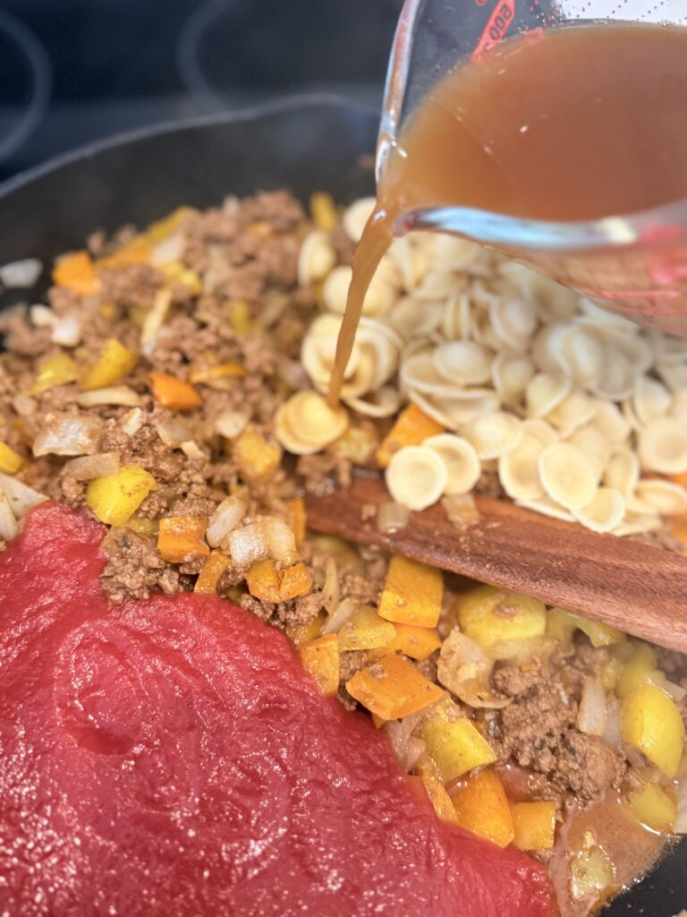 Ground beef with taco seasoning on top of it, red and yellow, diced bell peppers, diced onion, dried pasta, and minced garlic cloves, with broth being pour over top- all in a cast-iron pan on the stove-top.