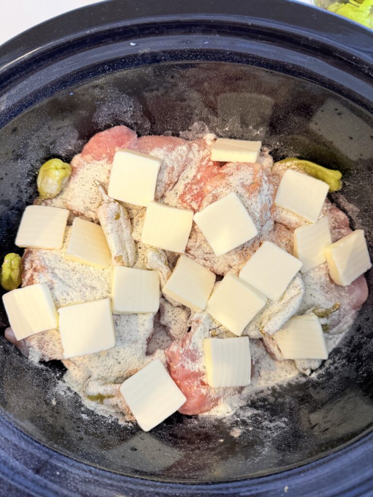 Chicken thighs, pepper, Cheney peppers, seasoning, packs, and tabs of unsalted butter, all layered in a slow cooker on the counter