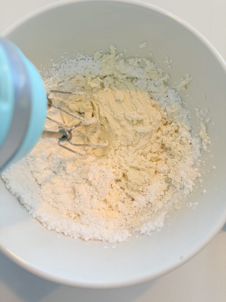 An electric hand mixer, mixing powdered sugar into butter and coconut extract and salt in a white mixing bowl on the counter.