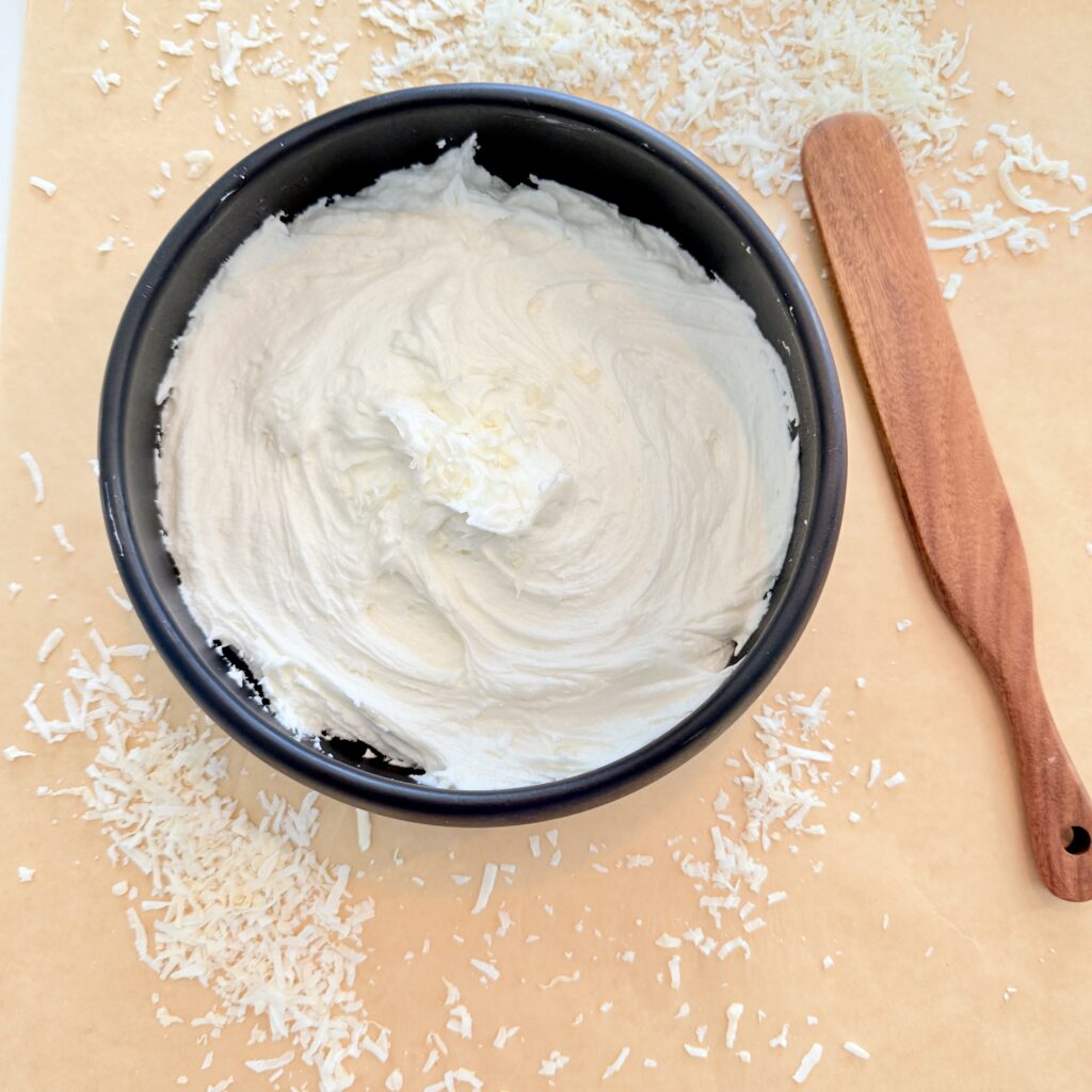 Coconut buttercream and a black bowl surrounded by coconut flakes and a brown spatula on brown parchment paper on the counter.