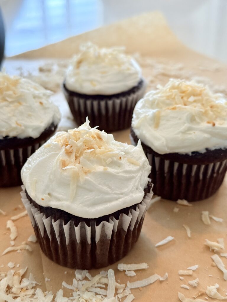 Chocolate cupcakes with coconut buttercream with toasted pieces of coconut on top on a brown parchment paper on the counter.