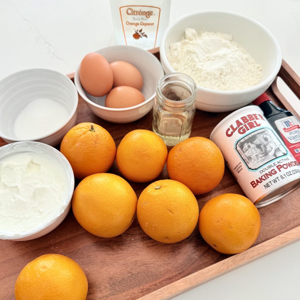 Orange Lecure on the counter, yogurt, plain Greek yogurt and a small white bowl on a brown cherry beside sugar and a small white bowl on the brown tray, three eggs and a small white bowl on the brown tray flower and a white bowl on the brown tray, vegetable oil in a glass jar on the brown tray, baking powder, and vanilla extract, along with oranges on the brown tray on the counter.