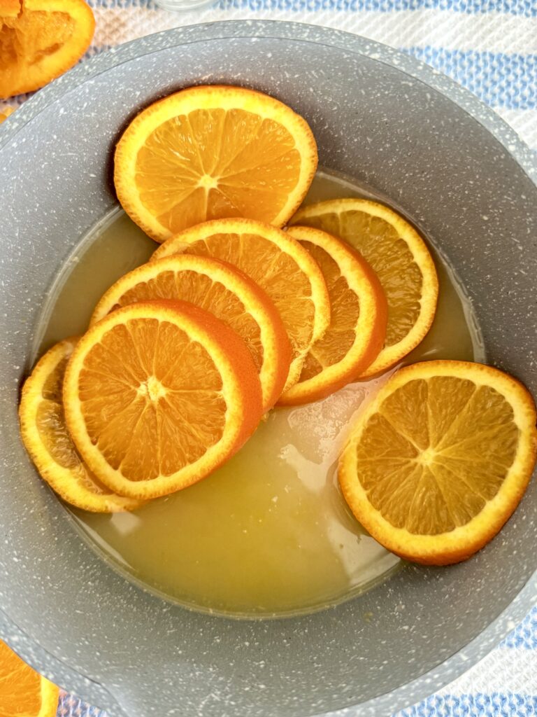 Slices of oranges, sugar, and orange juice in a pot. On the counter.