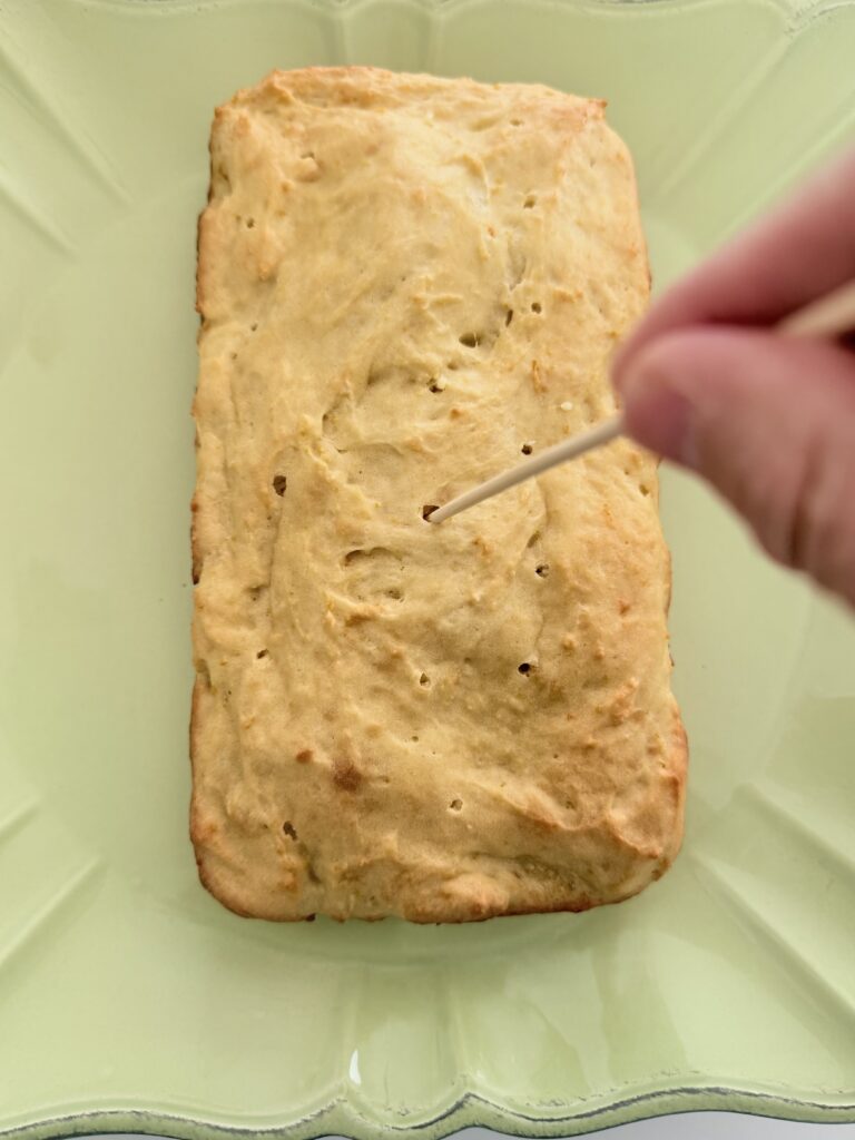 Using a wooden skewer to poke holes into the cake loaf on a green platter on the counter.