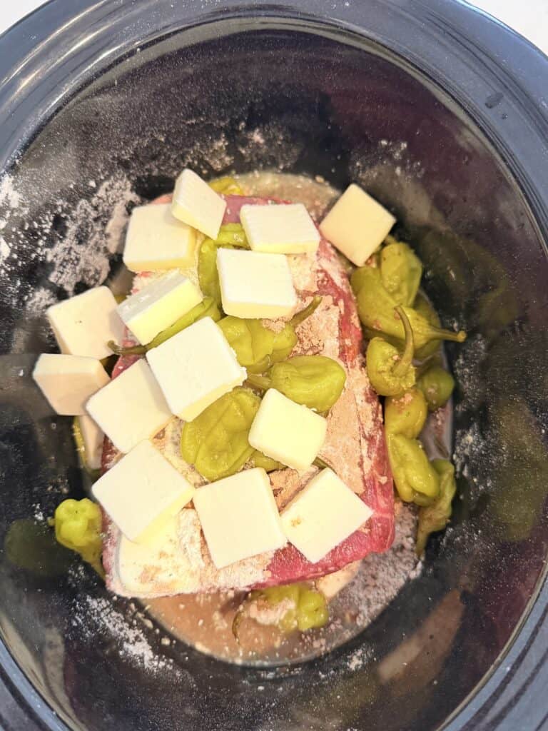 In a crockpot is the chuck roast, peppers and juice, packets of ranch seasoning, and gravy packet, unsalted tabs of butter on top of the meat in the crockpot on the counter.