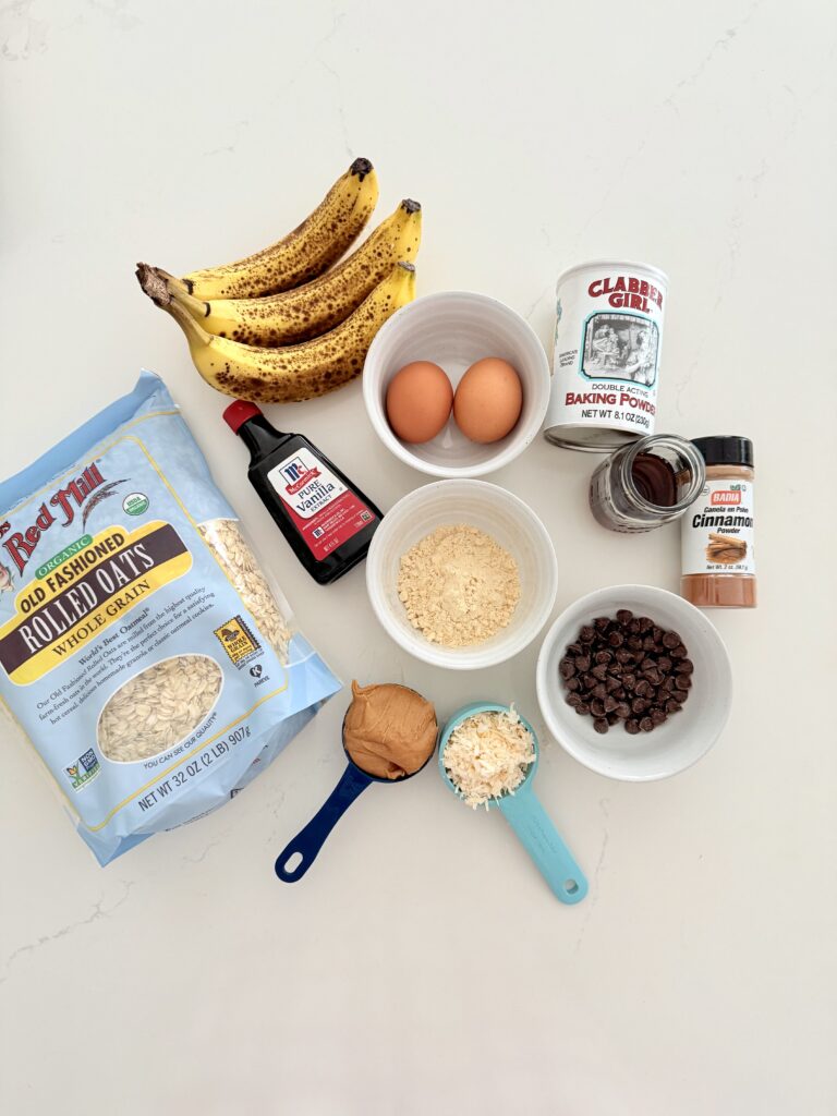 Write, bananas, rolled oats, maple syrup, eggs, vanilla extract, coconut flakes, protein powder, cinnamon, baking powder, semi sweet, chocolate chips, all on the counter. Milk is absent from the photo.