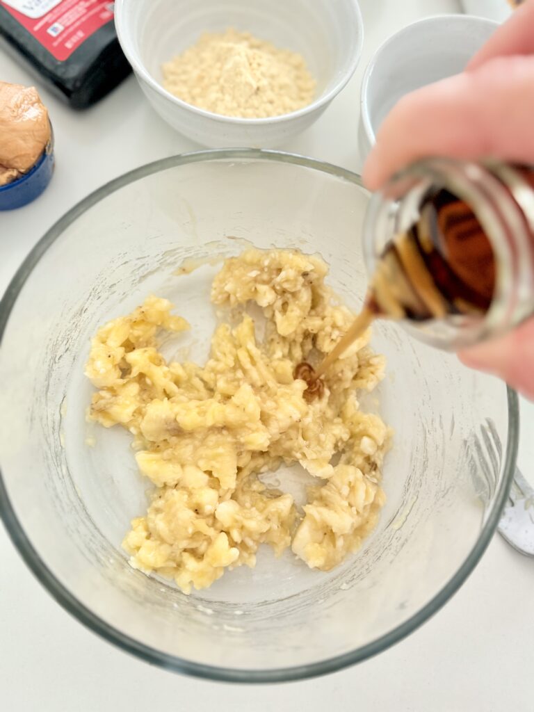 Pouring maple syrup into a glass bowl with mashed bananas all on the counter.