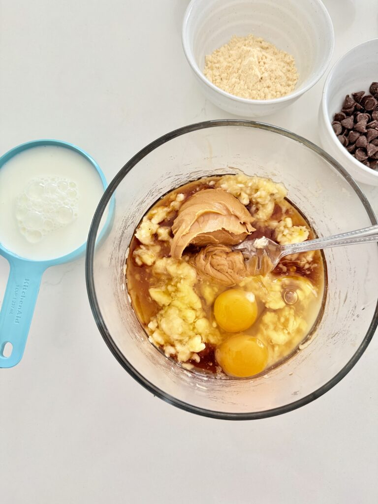 Mashed banana with wet ingredients in a glass bowl on the counter.