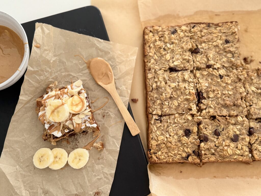 Chocolate chip banana breakfast bake, cut into squares to the right of a black cutting board with one piece of the chocolate chip banana breakfast bag drizzled with one peanut butter slices of banana and yogurt and slices of almonds all on the counter.