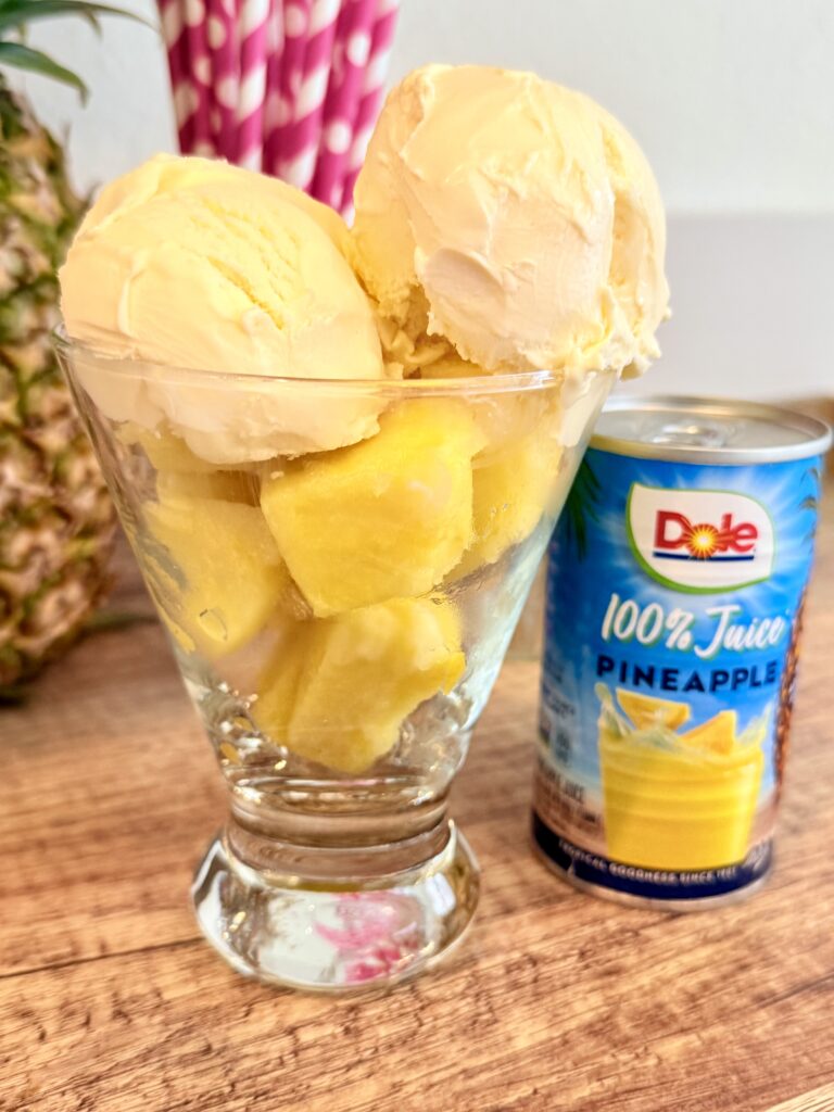  a glass of frozen pineapple chunks and two scoops of vanilla ice cream on a brown tray on the counter beside the glass with the frozen pineapple chunks and the ice cream scoops is a can of 100% pineapple juice. On the brown tray on the counter.