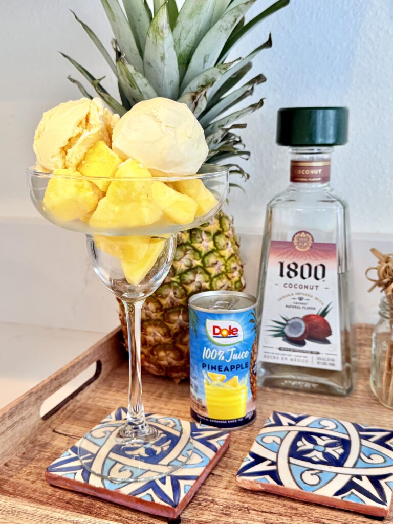 Vanilla ice cream, pineapple juice, frozen pineapple chunks, coconut tequila on a brown tray on the counter. The frozen pineapple trunks and the scoops of ice cream are in a margarita glass. In the background is a pineapple on a brown tray all on the counter.