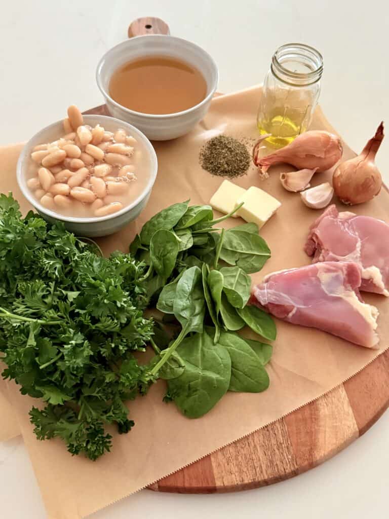 Olive oil in a glass jar, tabs of butter, herb, Province seasoning, shallots, garlic cloves, two pieces of skinless, boneless chicken thighs, a handful of fresh spinach and a handful of fresh parsley, white beans in a white bowl, and chicken broth and white bowl all on brown parchment paper on a brown cutting board on the counter.