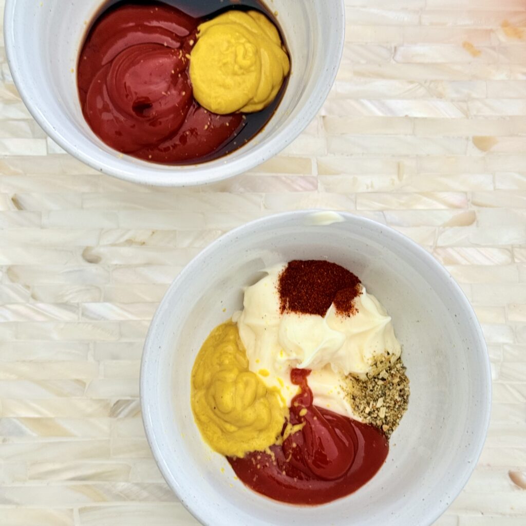 Mustard, ketchup, mayo, and spices are in a small white bowl on a tray on the counter. Beside it is another small white bowl that has the mustard, ketchup, and Worcestershire sauce on a tray.