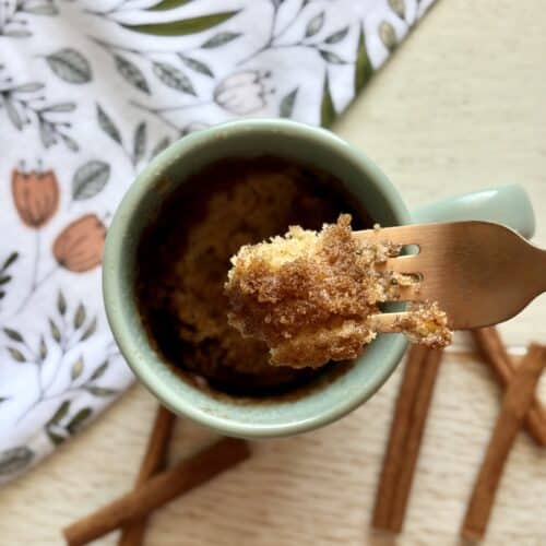 Close-up of a mug with cinnamon streusel muffin-in-a-mug on a wooden table, with cinnamon sticks and a floral cloth in the background.
