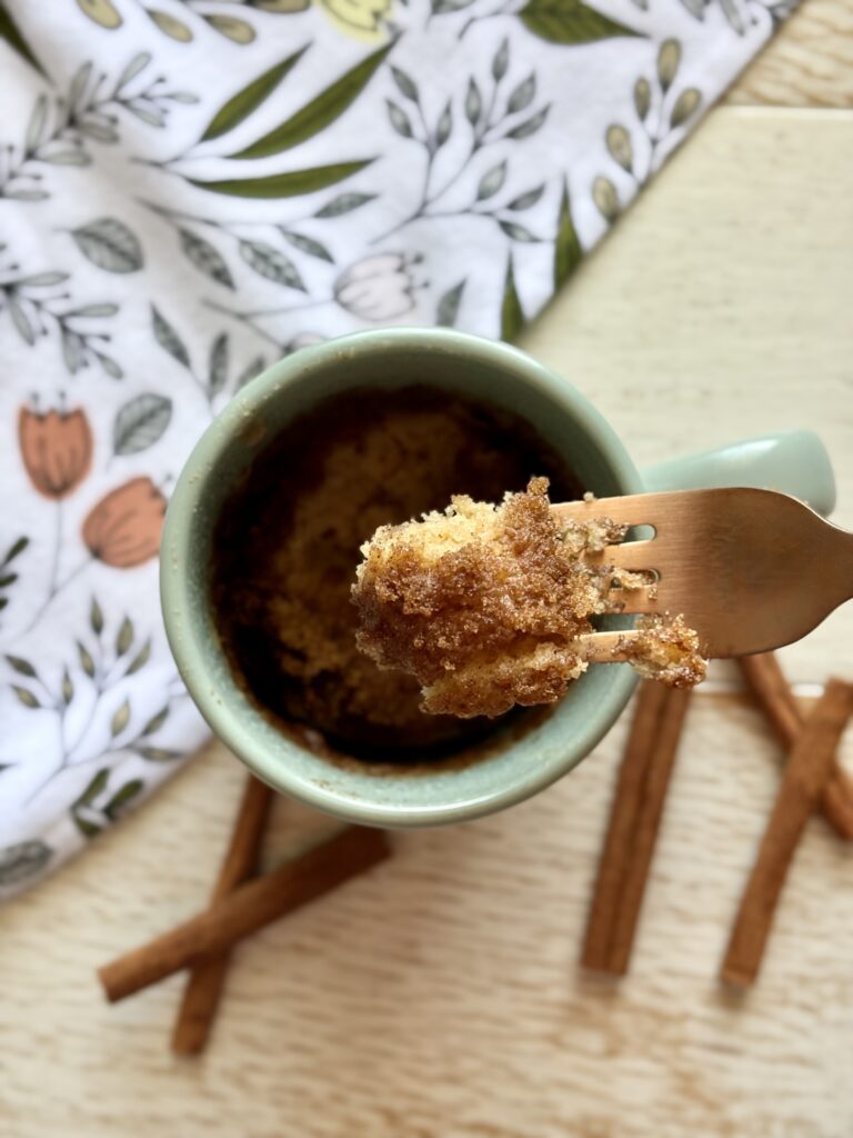 Close-up of a mug with cinnamon streusel muffin-in-a-mug on a wooden table, with cinnamon sticks and a floral cloth in the background.