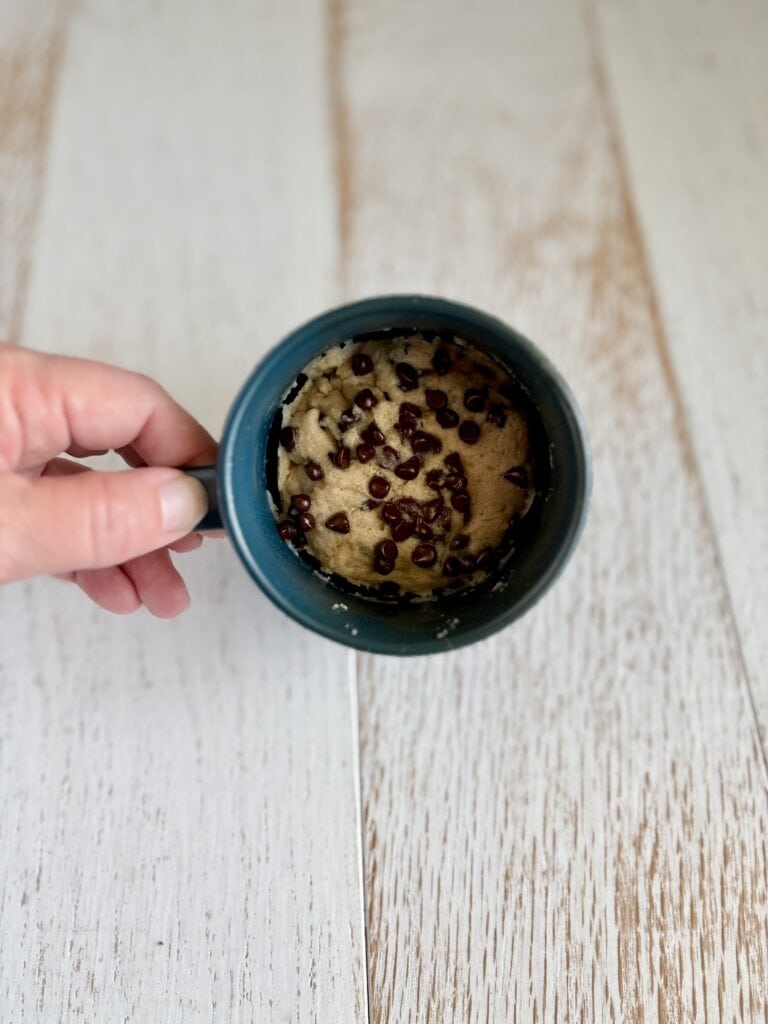 Close-up of a chocolate chip muffin in a mug, viewed from above, on a light wooden surface.