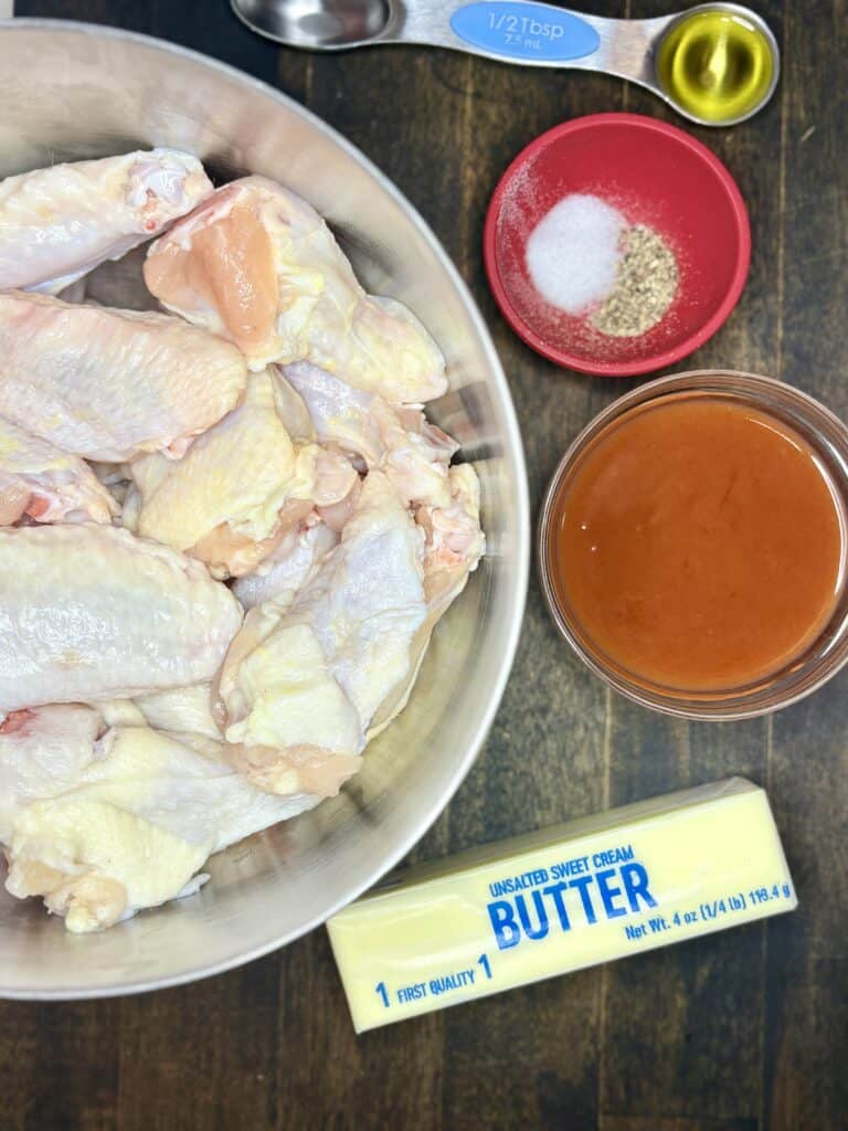 raw chicken wings in a bowl next to bowls of hot sauce and seasonings, a stick of butter, and a spoon of oil