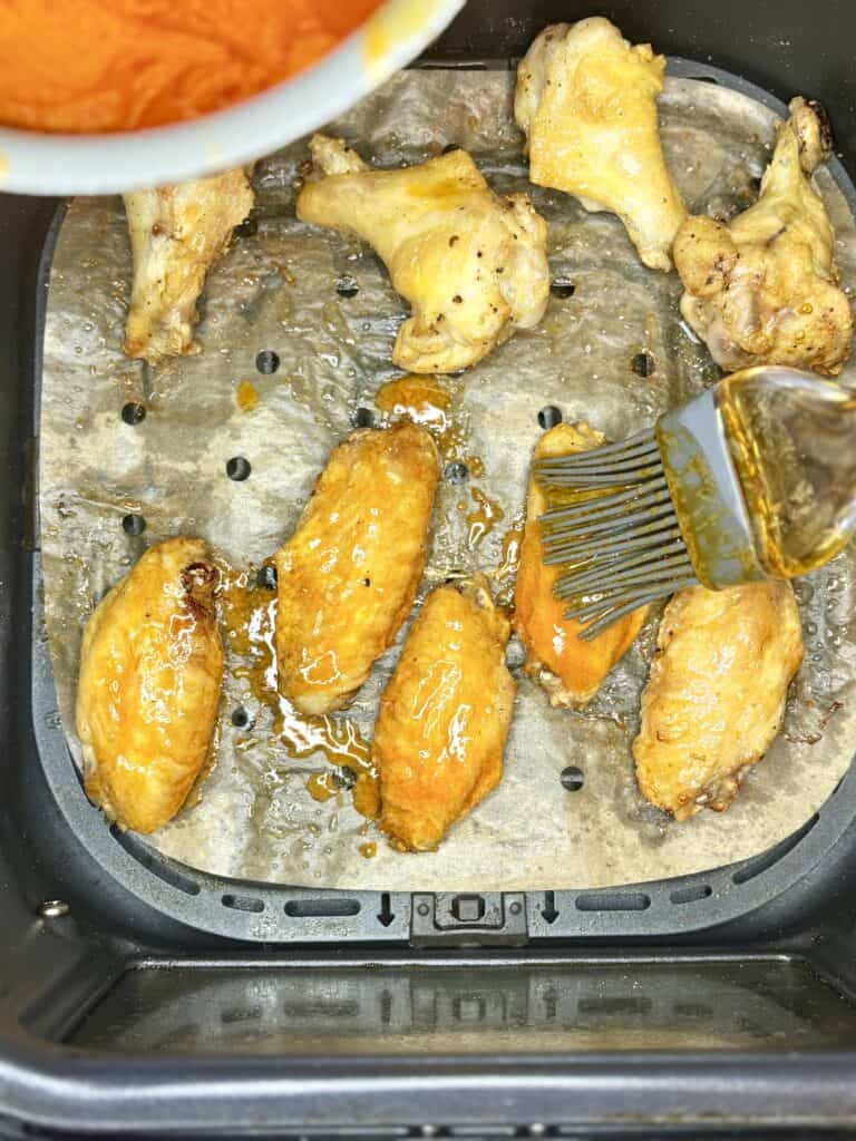 chicken wings in an air fryer basket being brushed with butter-hot sauce