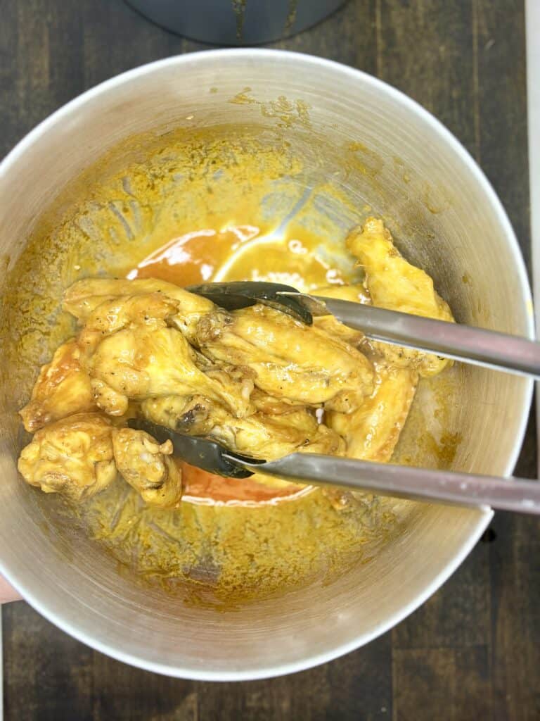 cooked chicken wings being stirred with tongs in a bowl with butter-hot sauce 