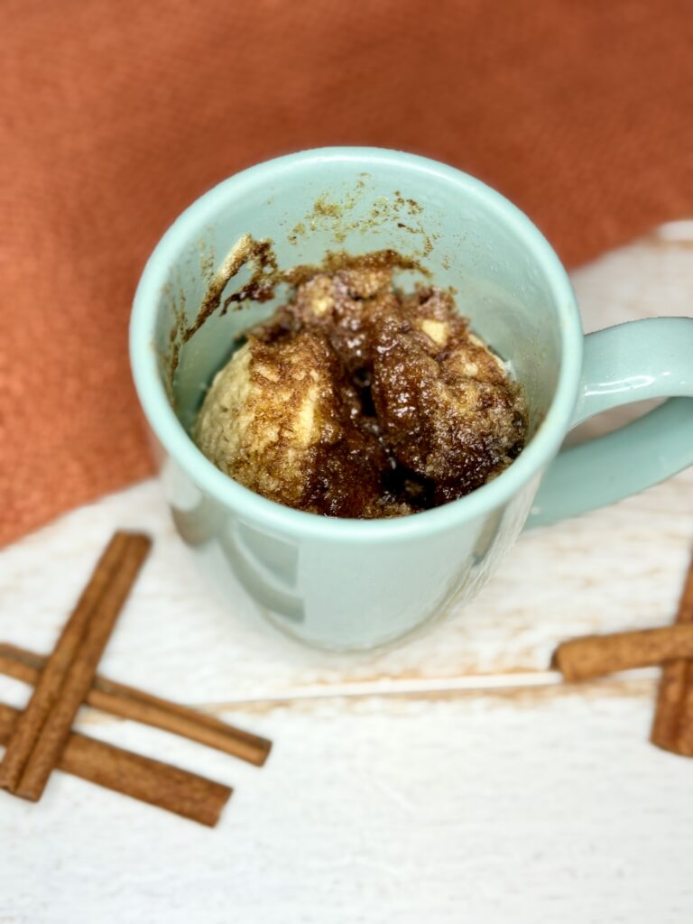 a mug with cooked cinnamon streusel muffin on a counter next to cinnamon sticks