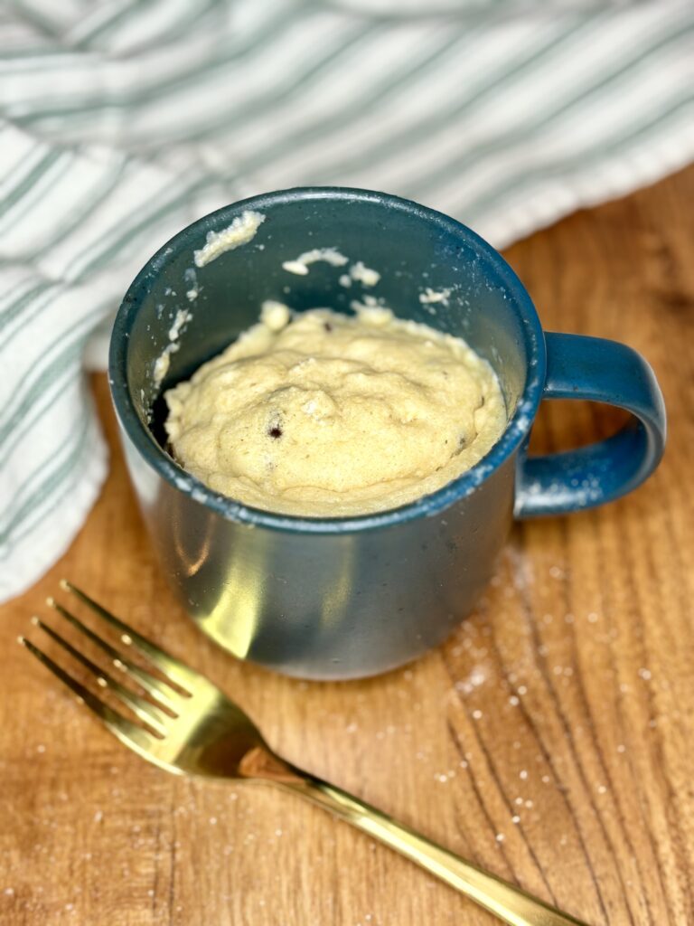 a cooked chocolate chip muffin in a mug next to a fork and towel on a cutting board 