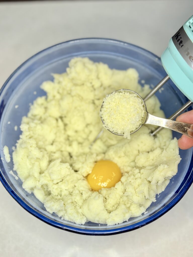 mashed potatoes in a bowl with an egg yolk and a tablespoon of parmesan cheese about to be beaten with a hand mixer