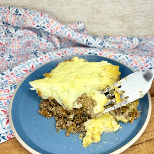 a fork cutting into a slice of shepherd's pie on a plate on a cutting board next to a kitchen towel