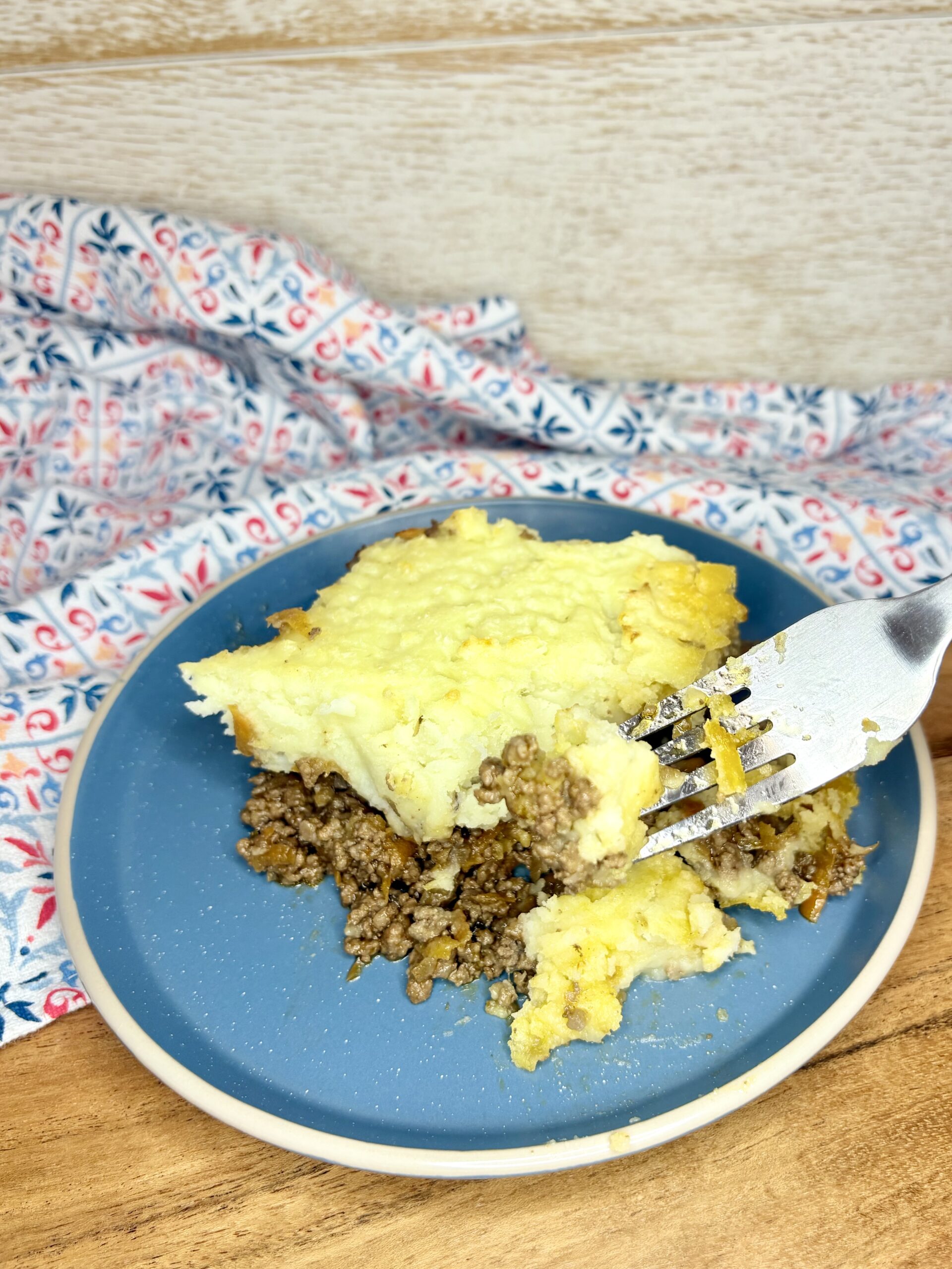 a fork cutting into a slice of shepherd's pie on a plate on a cutting board next to a kitchen towel