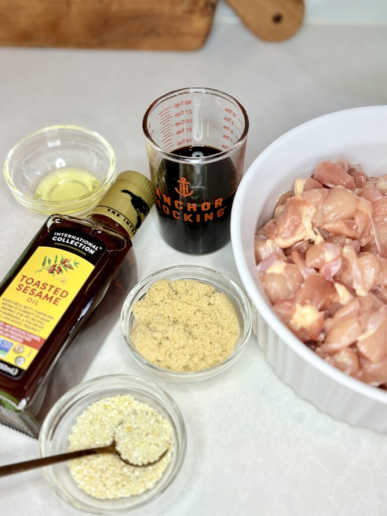 raw chicken in a bowl next to a measuring cup of soy sauce, bowls of brown sugar, oil, and sesame seeds, and a bottle of sesame oil