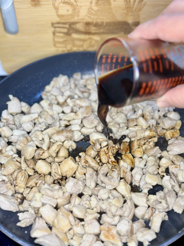 soy sauce being poured over chicken sautéing in a pan