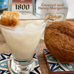 Coconut and Honey Margarita served in a clear glass with ice, garnished with a piece of honeycomb, on decorative tiles with a whole coconut and bottle of coconut honey in the background.