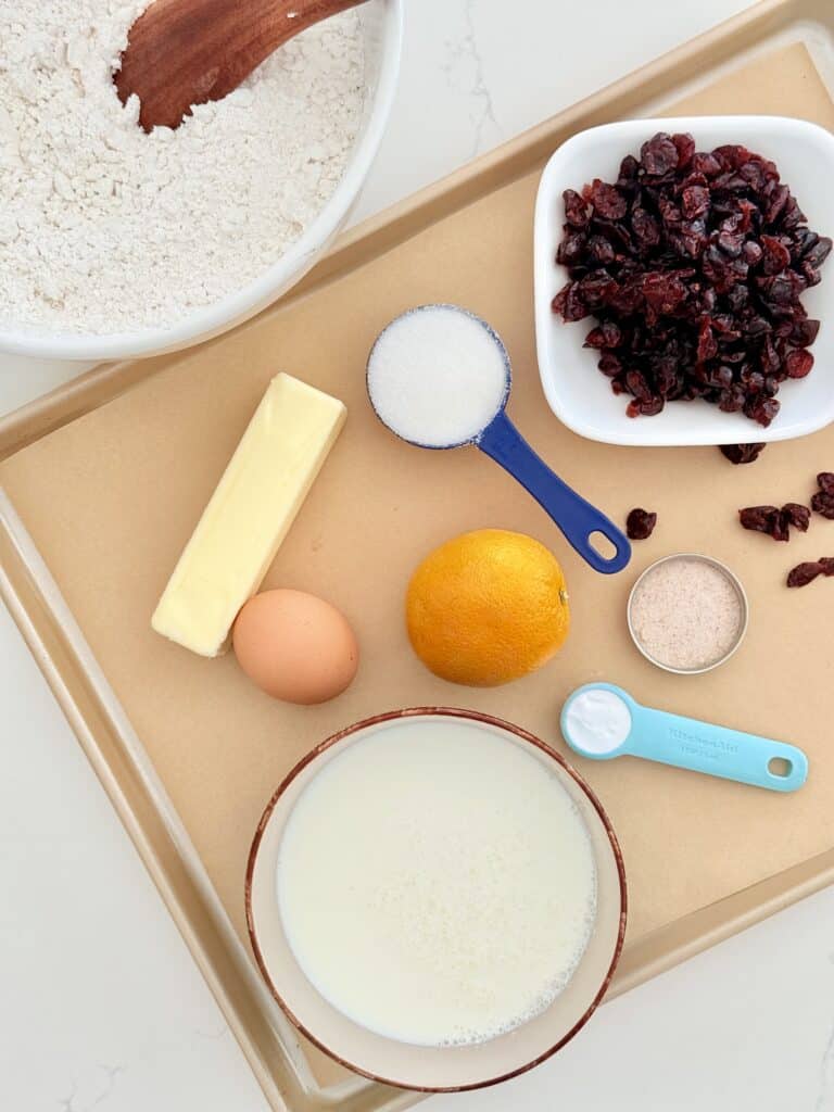 On the counter is large mixing bowl that is white with white flour and a brown one spoon in it next to it is a parchment paper lined baking sheet that is gold with a stick of unsalted butter, an egg, and medium size orange, buttermilk, and a white bowl, salt in a dish, dried cranberries and white dish sugar in a blue measuring cup, and baking soda in a light blue measuring cup all on the baking sheet that aligned with parchment paper on a white counter.