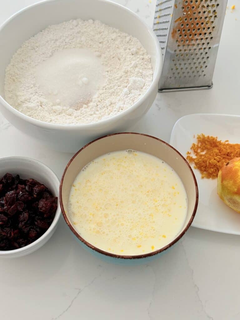 The dry ingredients in a white mixing bowl on the white counter, the wet ingredients in a blue bowl on the white counter, a cheese grater that is gray on the counter, the zest of a medium orange on a white plate with the remaining orange on the white plate beside it and dried cranberries in a small white bowl on the counter.