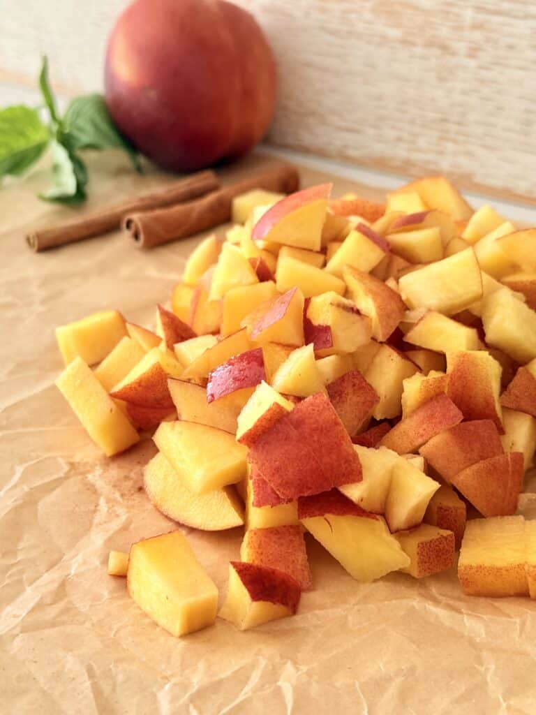 Bite-size cut pieces of nectarine on parchment paper on the counter with cinnamon sticks and a peach and basil leaves in the background. All on the counter.