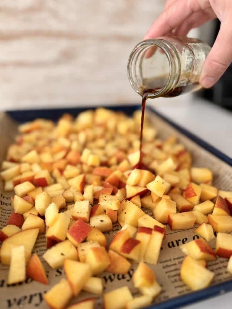 Drizzling bite-size pieces of nectarine with olive oil, balsamic vinegar, honey, cinnamon salt, and pepper mixture, all on a parchment paper lined blue baking sheet on the counter.