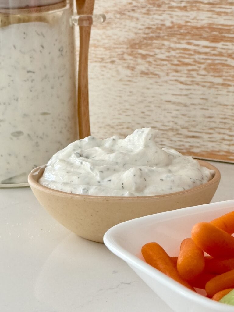 Homemade ranch in a sealed container in front of that homemade ranch and a small bowl and then in front of that in a small white bowl or cut up carrots and celery all on the countertop.