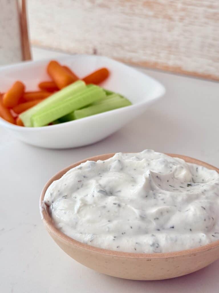 Homemade ranch dressing in a small wooden bowl on the counter with cut up celery and carrots in the background and white bowl on the counter.