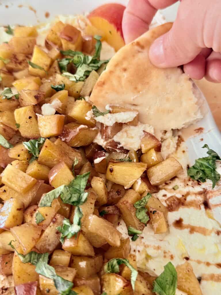 Using a piece of bread to pick up the whipped feta with roasted nectarines on a white platter on parchment paper on the counter.