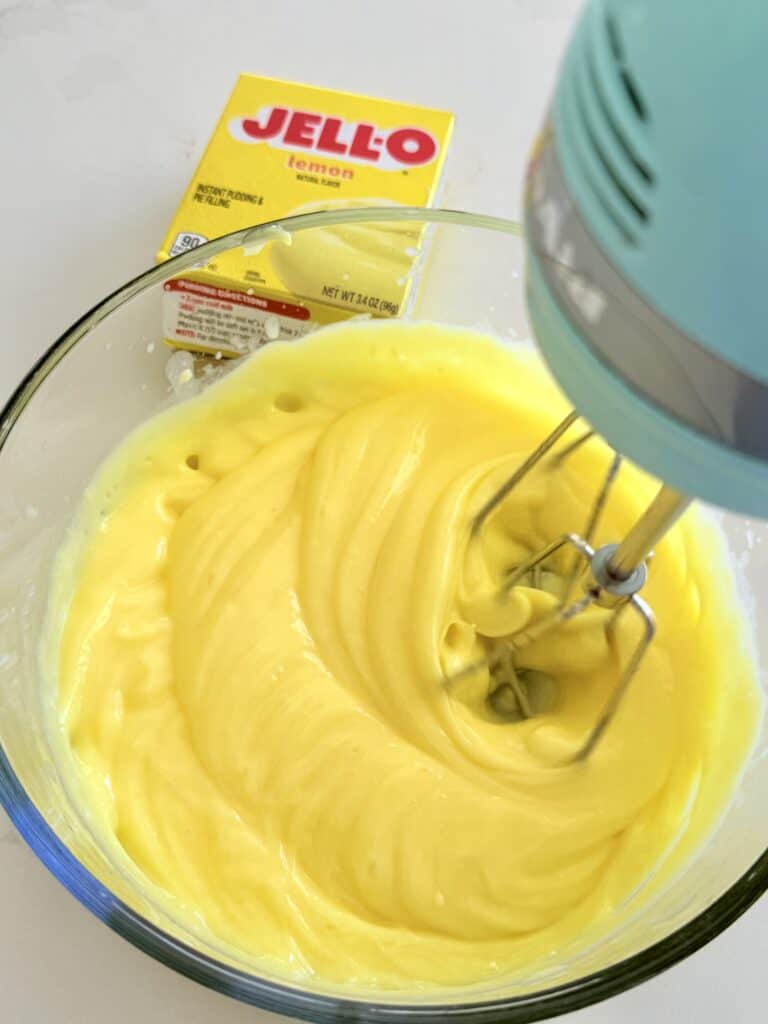 An electric hand and mixer mixes milk and Jell-O lemon pudding in a clear bowl on a white countertop.