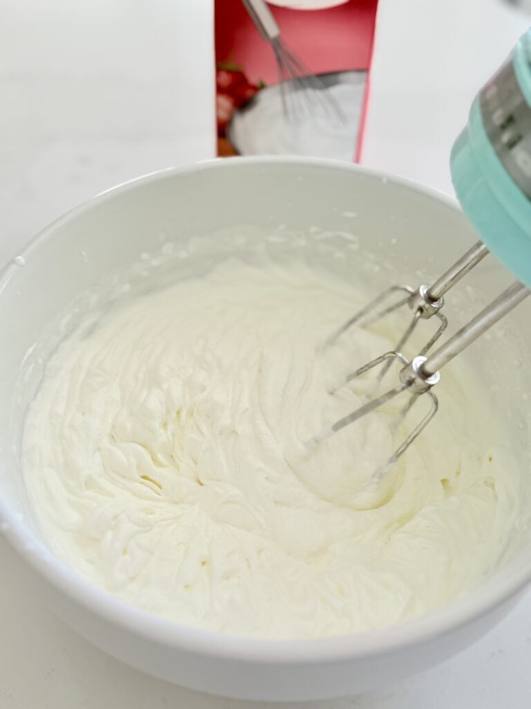 An electric mixer mixes heavy whipping cream on the counter in a white mixing bowl.