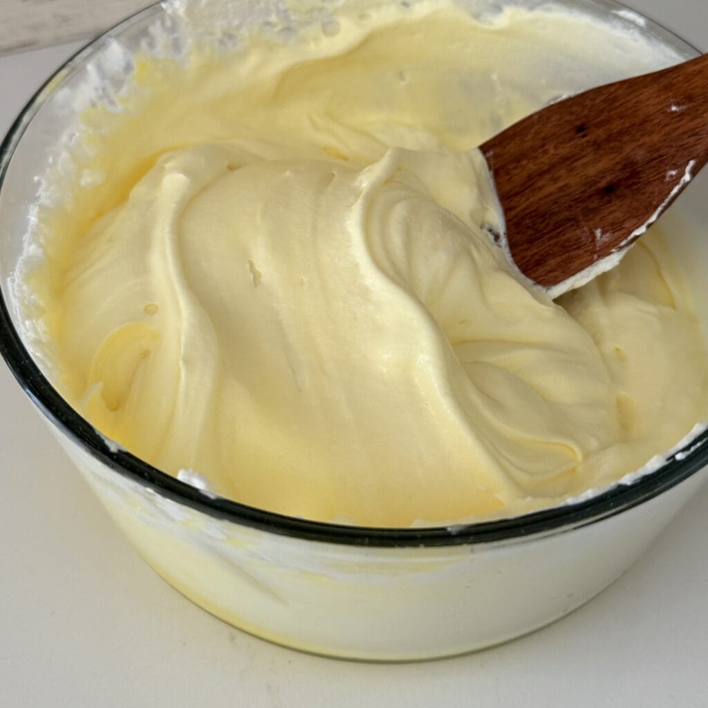 Folding together the lemon pudding and the whipping cream in a glass bowl with a wooden spoon on the counter.