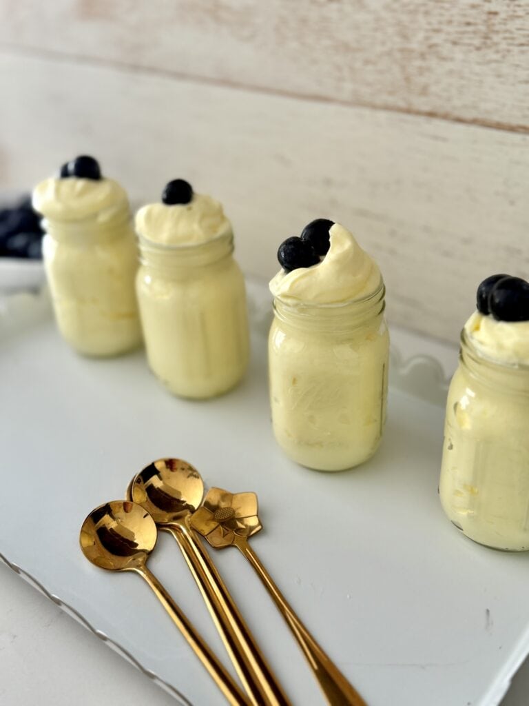 Simple lemon pudding, dessert in four glass small jars on a white tray with blueberries on top and gold spoons in front of the tray all on the counter.